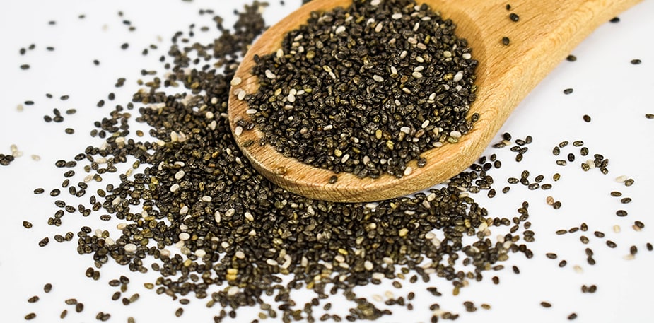 Een houten lepel met chia zaadjes erop, de chia zaadjes liggen er ook naast op een witte ondergrond